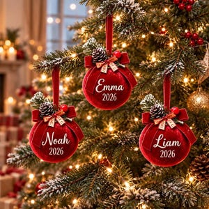 May include: Three round red velvet Christmas ornaments hanging from a tree. Each ornament features a red velvet ribbon, a small pinecone, and red berries. The names Emma, Noah, and Liam are printed in white script with the year 2026 below each name.