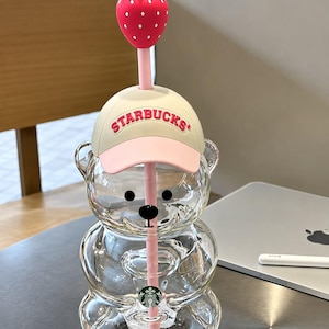 May include: A clear glass bear-shaped tumbler featuring a cream and pink baseball cap with the text STARBUCKS in pink lettering. A pink straw extends through the cap, topped with a red strawberry with white dots. A small green Starbucks logo is visible.