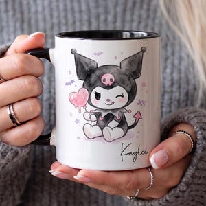 Puede incluir: Una taza de cerámica blanca con asa y borde interior negros, que presenta una ilustración estilo acuarela de un personaje de dibujos animados en blanco y negro con un emblema de calavera rosa, sosteniendo un globo de corazón rosa y el nombre Kaylee.