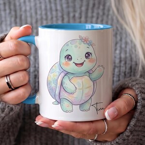 May include: A white ceramic mug with a bright blue handle and interior rim. The mug features a colourful illustration of a smiling baby turtle with a pastel-coloured shell, a small flower on its head, and a signature near the bottom right corner.
