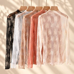 May include: A collection of five long-sleeve shirts made of sheer, floral-patterned lace fabric. The shirts are displayed on wooden hangers and come in black, white, beige, coral, and light pink, each featuring a high neckline and long sleeves.