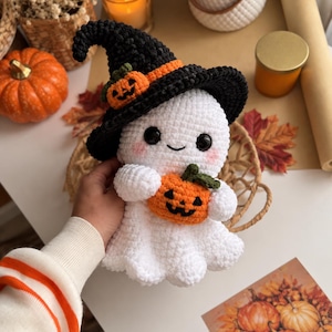 May include: A white crocheted ghost plush toy wearing a black witch hat with an orange band and a small pumpkin accent. The ghost has black safety eyes, a stitched smile, pink cheeks, and is holding a small orange crocheted pumpkin.