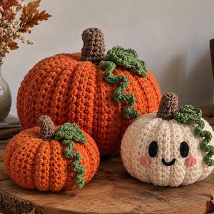 May include: Three crocheted pumpkins in varying sizes. One large orange pumpkin, one small orange pumpkin, and one small cream-coloured pumpkin with a smiling face featuring black oval eyes, a curved mouth, and pink circular cheeks. Each has a brown stem.