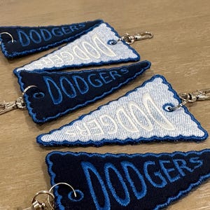 May include: Five triangular pennant keychains featuring the word DODGERS embroidered in blue or white thread. Three pennants are dark navy blue with blue embroidery, and two are light blue denim with white embroidery, each with a metal clasp.