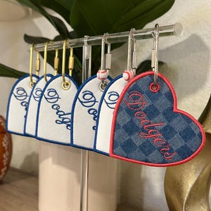 May include: A collection of heart-shaped keychains hanging from a clear stand. Several white fabric hearts feature blue embroidered text, while one blue checkered fabric heart features red embroidered text reading Dodgers.