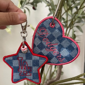 May include: Two fabric keychains featuring a blue and grey checkered denim pattern with bright pink stitched edges. One keychain is star-shaped with a red embroidered LA logo, and the other is heart-shaped with the word Dodgers in pink script.