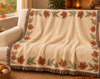May include: A cream-colored woven throw blanket featuring a decorative border of orange maple leaves and green pine sprigs. The blanket has a textured surface and a fringed edge, draped over a sofa to display the repeating botanical pattern.