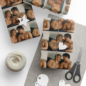 May include: Wrapping paper featuring a repeating photographic print of two shirtless men lying together. The paper is tied with brown twine and decorated with white star and heart-shaped gift tags, accompanied by a ball of white yarn and scissors.
