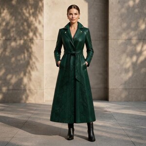 Könnte beinhalten: Ein langer, dunkelgrüner Trenchcoat mit texturiertem Schlangenledermuster, breitem Reverskragen, goldfarbenen Knöpfen und einem passenden Bindegürtel in der Taille. Der Mantel hat lange Ärmel, einen bodenlangen Rock und wird mit schwarzen Stiefeletten kombiniert.
