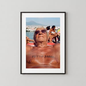 May include: A framed photographic print featuring a sun-tanned individual wearing brown sunglasses while reclining on a beach chair. The person has the words TUTTO PASSA tattooed in black ink across their chest, with a coastal background.