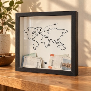 May include: A black shadow box frame containing a white background with a continuous black line drawing of a world map. The map features a dotted flight path, a small airplane icon, and a hot air balloon. Two stacks of paper boarding passes sit inside.