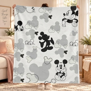 May include: A white fleece blanket featuring a repeating pattern of black and grey Mickey Mouse silhouettes and illustrations. The design includes various poses of the character interspersed with solid grey and outlined Mickey head shapes.