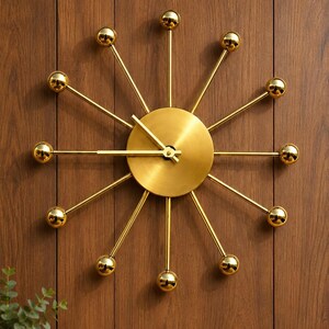 May include: A gold sunburst wall clock featuring a circular center dial with matching hour and minute hands. Twelve thin metal spokes extend outward from the center, each tipped with a polished gold sphere, creating a star-like radial design.