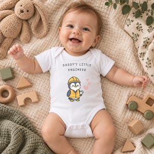 Op de afbeelding: Een wit babyrompertje met korte mouwen en de tekst DADDY'S LITTLE ENGINEER in zwarte letters boven een afbeelding van een pinguïn met een gele veiligheidshelm en oranje veiligheidsvest, die een blauwdruk vasthoudt, met drie kleine roze hartjes.