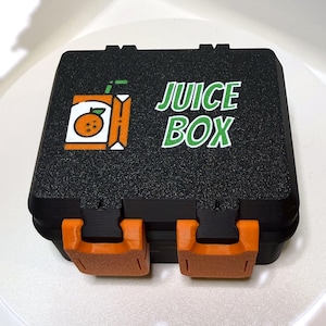 May include: A black rectangular storage case with two orange latches on the front. The lid features a graphic of an orange juice box and the white text JUICE BOX. The case has a textured finish and a hinged design for secure closure.