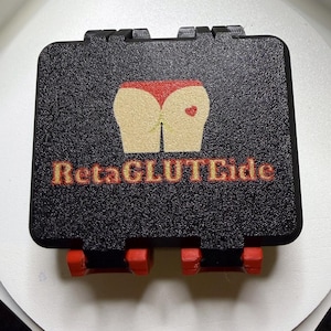 May include: A rectangular black storage case with a textured surface featuring a graphic of a pair of buttocks wearing red underwear with a small red heart. The text RetaGLUTeide is printed in orange and yellow below the graphic with red latches.