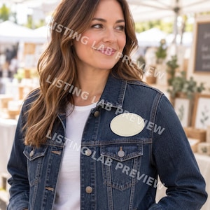 May include: A dark blue denim jacket featuring a plain, oval-shaped cream-colored patch sewn onto the left chest area. The jacket has a classic collar, metal buttons down the front, and two buttoned chest pockets. A white t-shirt is worn underneath.