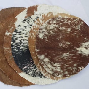 Può includere: Una collezione di cinque tappeti circolari in pelle animale sovrapposti l'uno sull'altro. I tappeti presentano vari motivi e colori naturali, tra cui texture di pelliccia color marrone chiaro, marrone e bianco maculato e nero e bianco.