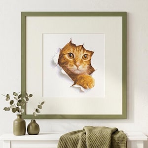 May include: A framed cross-stitch artwork featuring an orange tabby cat peeking through a torn white paper effect. The cat has bright amber eyes and one paw extended forward. The square frame is olive green with a wide off-white matting border.