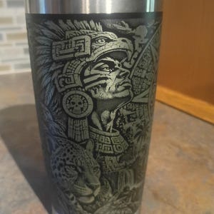May include: A stainless steel tumbler with a black matte finish featuring a detailed laser-engraved design of an Aztec warrior wearing an ornate feathered headdress, a skull medallion, and a jaguar positioned in the foreground of the scene.