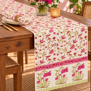 May include: A cream-coloured table runner featuring a repeating pink floral vine pattern with green stems. The runner has a distinct border with a horizontal band of pink flowers and a bottom edge finished with a vertical green and white striped trim.