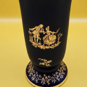 May include: A dark blue vase with a scalloped rim and a gold-coloured trim. The vase features gold-coloured illustrations of a couple in period clothing, floral garlands around the top, and delicate botanical patterns on the base and interior rim.
