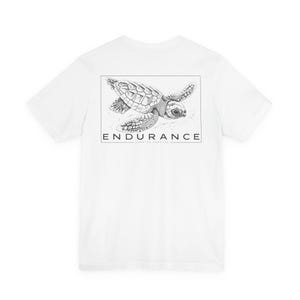 May include: A white short-sleeved t-shirt featuring a black and white line art illustration of a sea turtle inside a rectangular frame on the back. Below the turtle, the word ENDURANCE is printed in capital letters with Caretta Caretta below it.
