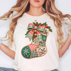 May include: A cream-colored t-shirt featuring a large graphic of a Christmas stocking. The stocking is designed with a patchwork pattern of green, red, and white plaid, gingham, and snowflake prints, topped with a large bow and pine needles.