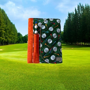 May include: A quilted fabric scorecard holder featuring a green background with a pattern of white golf balls and colorful tees. It includes a solid orange spine, a pen loop with a decorative pen, and a small grey tag labeled JEM.