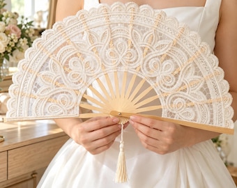 Puede incluir: Un abanico plegable de mano con intrincado tejido de encaje blanco, borde festoneado y un patrón floral detallado. El abanico está sostenido por varillas de madera clara e incluye una pequeña borla blanca a juego que cuelga del pivote central.