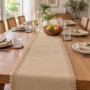 May include: A long, rectangular tan burlap table runner with frayed, decorative edges centered on a wooden dining table. The runner features a woven texture and is positioned beneath a wooden bowl filled with pears and apples, and a glass pitcher.
