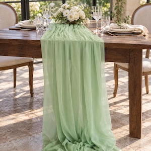 May include: A sage green chiffon table runner draped over a dark wooden dining table. The fabric features a soft, semi-sheer texture with vertical folds that cascade down the side of the table, creating a flowing and layered appearance.