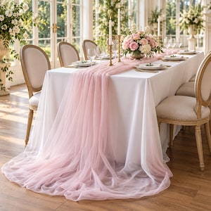 May include: A long, sheer, pale pink chiffon table runner draped over a white tablecloth on a rectangular dining table. The runner pools onto the floor, featuring a soft, translucent texture. The table is set with gold candlesticks and floral centerpieces.