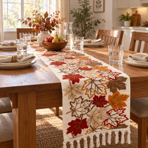 May include: A cream-colored fabric table runner featuring embroidered maple leaves in shades of red, orange, and brown. The runner is finished with decorative fringe tassels at the ends and is displayed on a wooden dining table with place settings.
