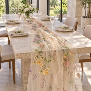 Könnte beinhalten: Ein transparenter, cremefarbener Tischläufer mit aufwendiger, mehrfarbiger Blumenstickerei in Gelb, Rosa, Blau und Grün. Der Läufer liegt auf einem langen Esstisch aus Stein, der mit weißen Tellern, Gläsern und einer Blumenvase gedeckt ist.