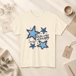 May include: A cream-coloured short-sleeve t-shirt featuring a graphic of blue stars with dark blue outlines and splatters. The text reads, "Each star is unique and shines brightly" and "eternal light" in a dark blue script font.