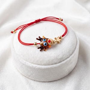 Può includere: Un bracciale in cordino rosso con un ciondolo a forma di renna in vetro con occhi blu e naso rosso, arricchito da una piccola stella dorata e perline dorate. Il bracciale presenta una chiusura scorrevole regolabile con perline dorate.