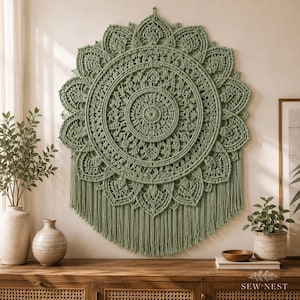 Könnte beinhalten: Ein salbeigrüner Makramee-Wandbehang mit kreisförmigem Mandala-Design, komplizierten Knotenmustern und einem Rand aus überlappenden Blütenblättern. Das Stück hat unten lange, gerade Fransen und ist mittig an einer Wand über einer Konsole platziert.