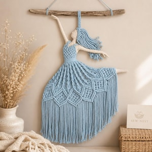 Könnte beinhalten: Ein Makramee-Wandbehang, der eine Figur in einem hellblauen Kleid zeigt, die an einem Stück Treibholz hängt. Die Figur hat cremefarbene Arme und einen Kopf, langes hellblaues Haar und ein Kleid mit komplizierten Knotenmustern und Fransen.