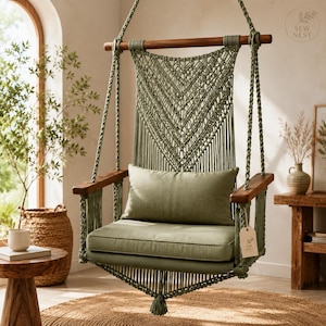 May include: A sage green macrame hanging chair featuring a wooden top bar and armrests, a V-shaped knotted backrest, and a matching rectangular seat cushion. A small tag with the text SEW NEST is attached to the side of the chair frame.