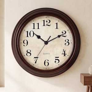 May include: A round wall clock with a dark brown frame and a cream-colored face featuring black Arabic numerals. The clock has black hour, minute, and second hands, with the word QUARTZ printed in black text below the center of the clock face.