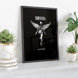 Könnte beinhalten: Ein schwarz gerahmter Kunstdruck mit dem Nirvana In Utero Albumcover-Motiv, das eine weiße geflügelte Figur auf schwarzem Grund zeigt. Der Text NIRVANA steht über der Figur und IN UTERO darunter in einer weißen Serifenschrift.