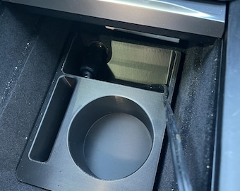 May include: A black plastic center console organizer insert for a vehicle, featuring a circular cup holder section and a rectangular storage slot. The insert is positioned within the car's console area, with a black power cable visible nearby.