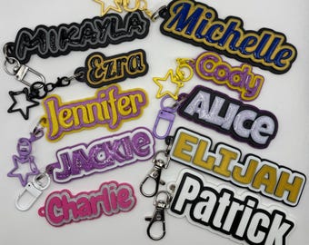May include: A collection of custom name keychains featuring various colors and glitter finishes. Each keychain displays a unique name including Mikayla, Ezra, Jennifer, Jackie, Charlie, Michelle, Cody, Alice, Elijah, and Patrick, attached to metal star-shaped clasps.