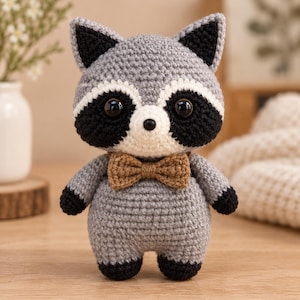 May include: A crocheted raccoon plush toy featuring a light grey body, black eye patches, black ears, and black paws. The raccoon has a white muzzle, a small black nose, large dark eyes, and a brown crocheted bow tie attached at the neck.