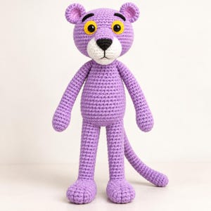May include: A crochet plush toy shaped like a panther with light purple yarn, a white muzzle, a black nose, and large yellow eyes with black pupils. The figure has rounded ears, long arms, and a long tail, standing upright on two feet.