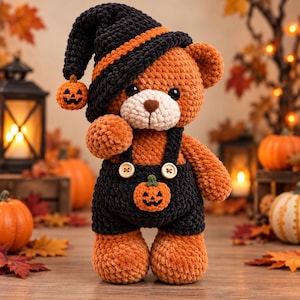 Puede incluir: Un oso de peluche tejido a ganchillo con hilo naranja suave, que lleva un peto negro con dos botones claros y un pequeño aplique de calabaza de Halloween. El oso tiene un gorro negro a juego con una raya naranja y una pequeña calabaza colgante.