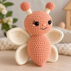 Puede incluir: Un juguete de peluche tejido a ganchillo con forma de abeja, con cuerpo y cabeza de color melocotón. Presenta dos antenas de color crema con puntas melocotón, cuatro alas color crema, ojos de plástico negro con pestañas bordadas y una sonrisa.