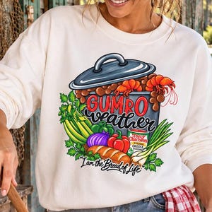 May include: A cream-colored long-sleeve sweatshirt featuring a graphic of a pot of gumbo with shrimp, vegetables, and the text "GUMBO weather" and "I am the Bread of Life" printed on the front, worn with a red and white checkered apron.