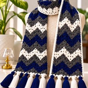May include: A crochet scarf featuring a chevron pattern in alternating stripes of navy blue, charcoal grey, and white. The scarf is finished with a white crocheted border and alternating navy blue and white tassels along the bottom edges.